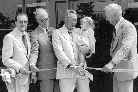 Four men stand together as one cuts the ribbon to open a building while also holding a little girl